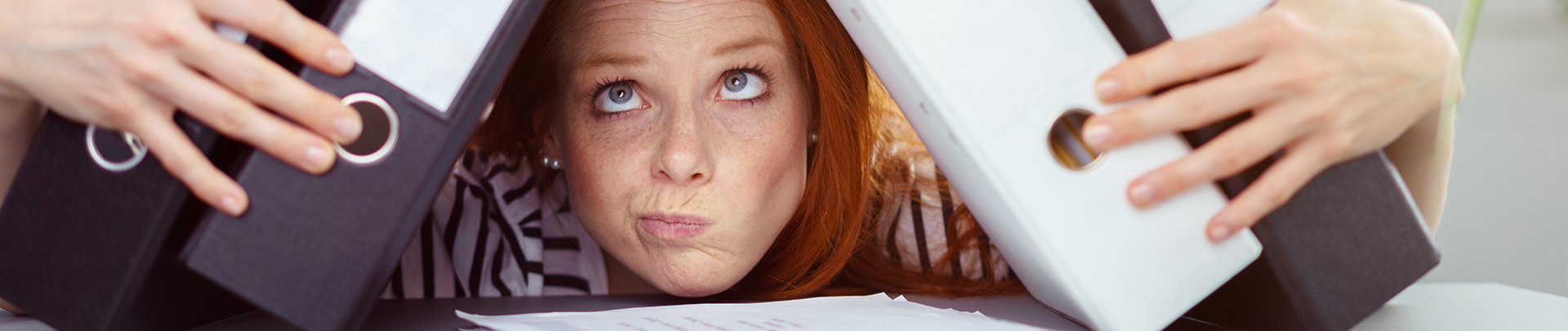 Burn-out; jonge vrouw met haar kin op een stapeltje papieren op haar bureau en houd ordners tegen haar hoofd aan